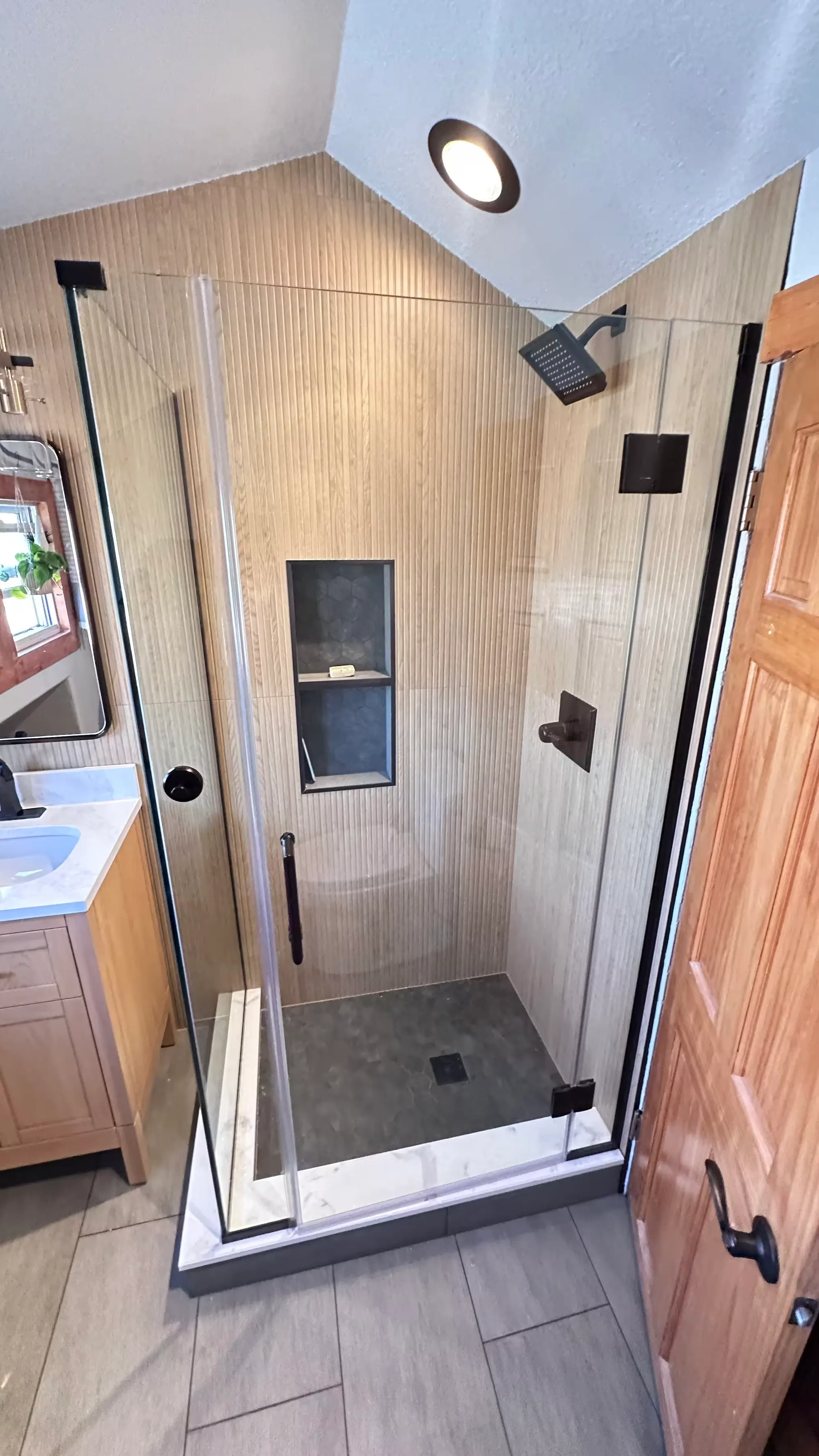 A glass-enclosed Denver shower with beige vertically ridged wall tile and a recessed niche stands beside a wood door.