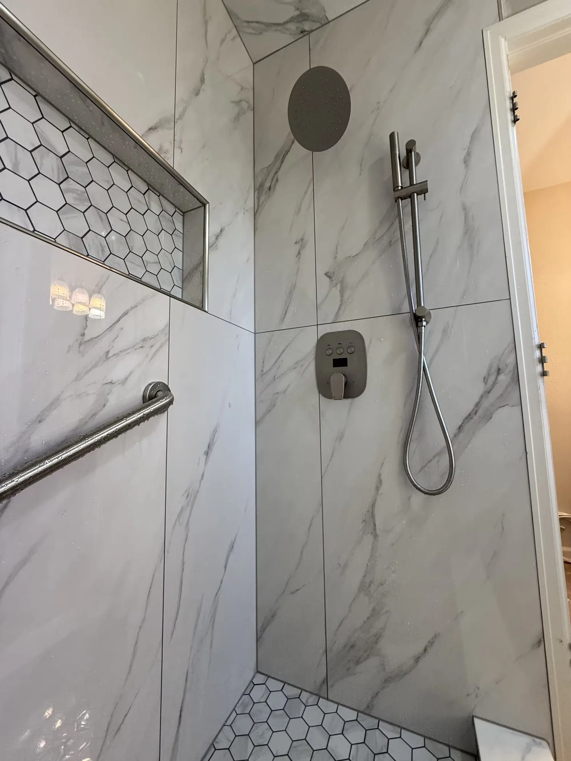 The fixture wall of a Littleton shower includes a round rainfall showerhead, a handheld shower, a grab bar, and a recessed niche.