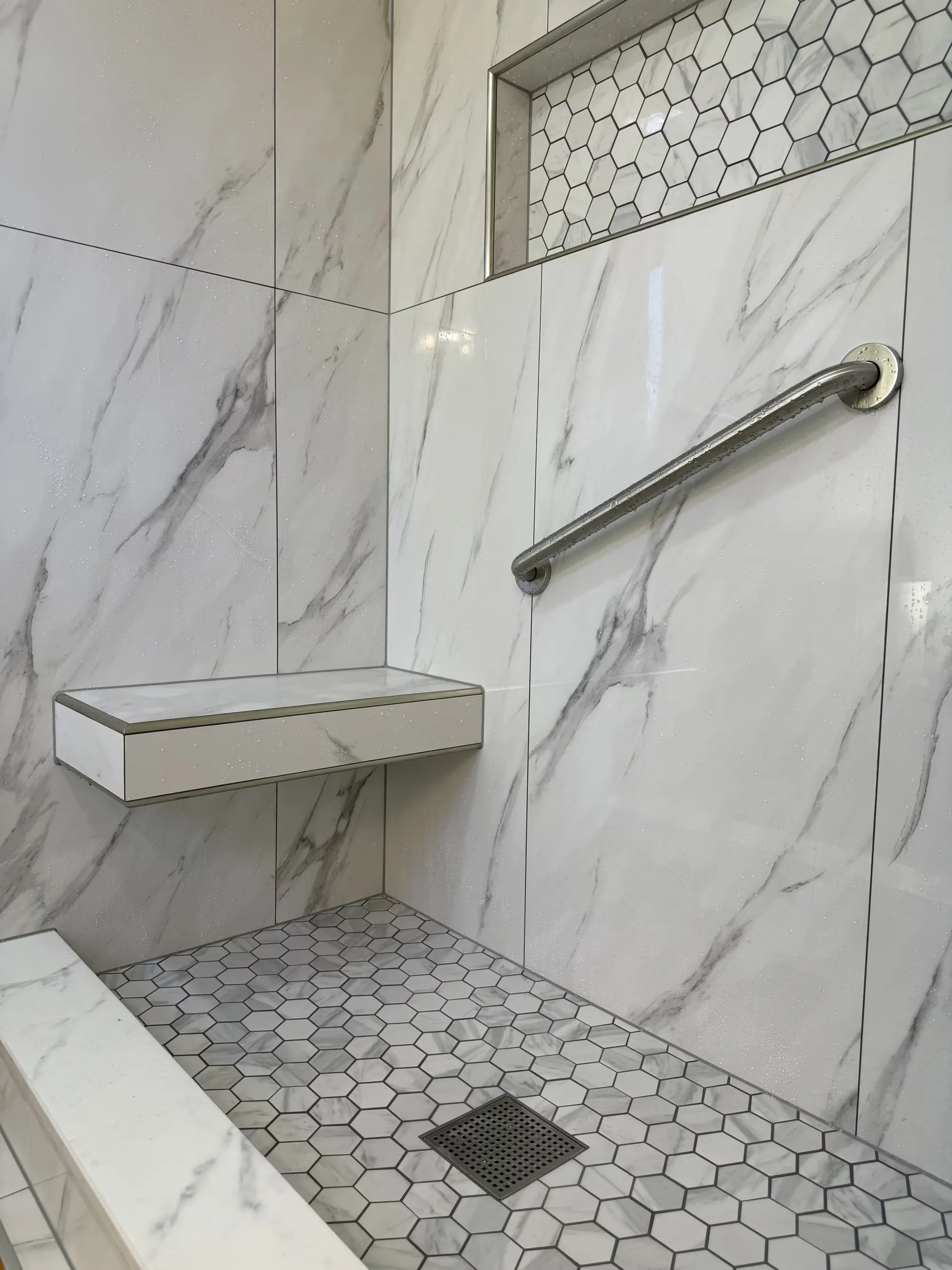 A Littleton shower corner shows a marble-look floating bench, a metal grab bar, and a hexagon-tile floor with a square drain.