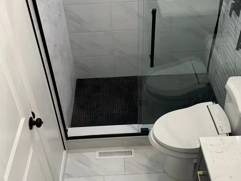 An Aurora bathroom shows a black-framed glass shower with light marble-look wall tile beside a white toilet and dark vanity.