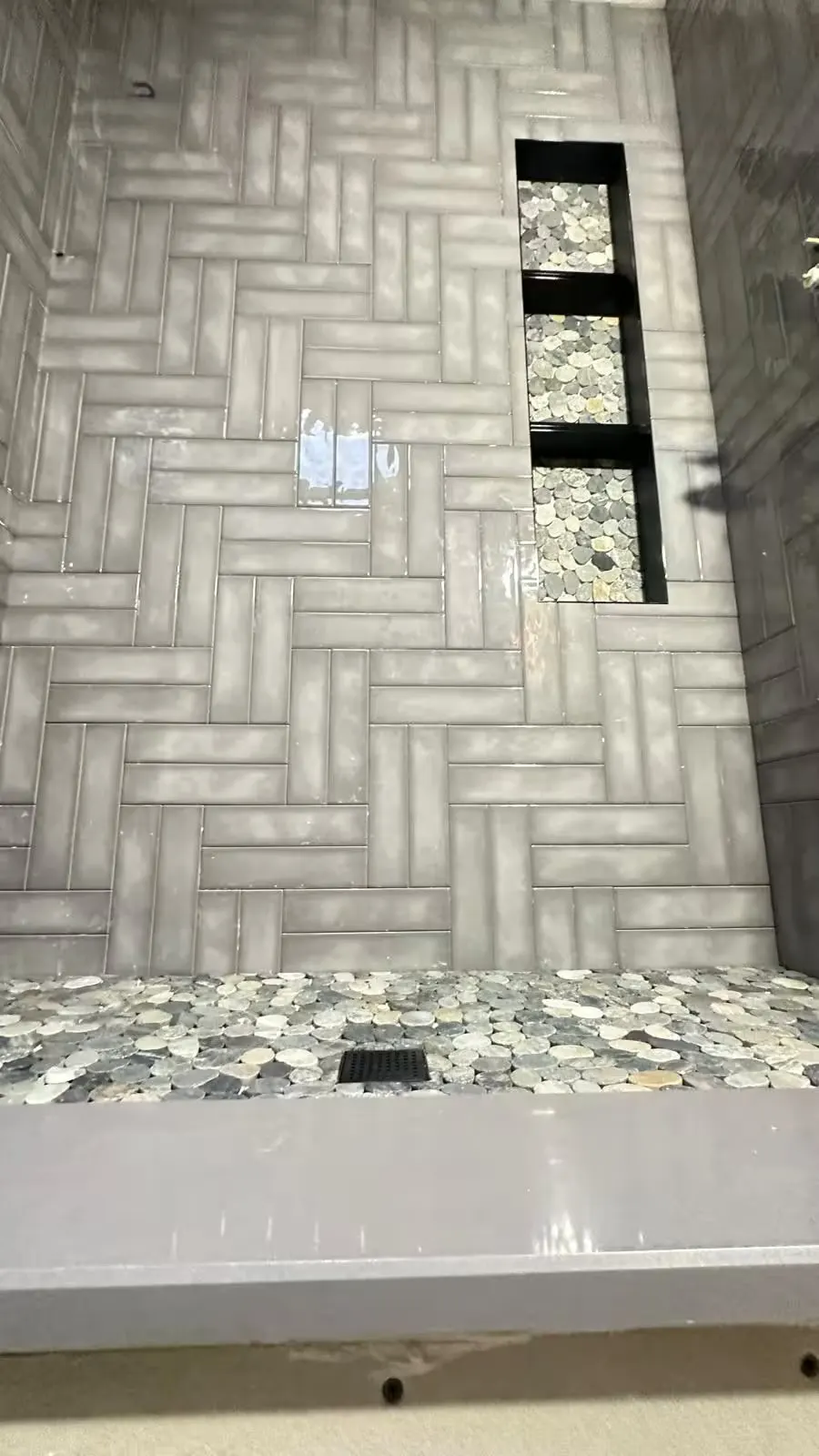 An Arvada shower has gray herringbone wall tile, a pebble-tile floor, and a tall recessed niche with three shelves.