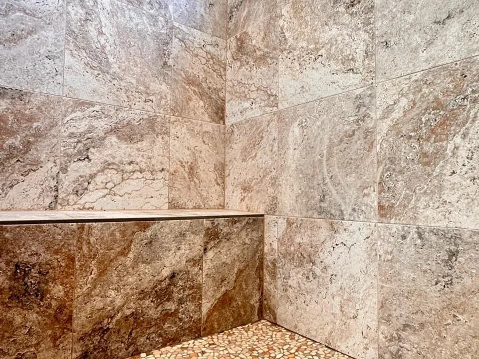 Tan wall tile and a pebble floor are visible in a finished Aurora shower.